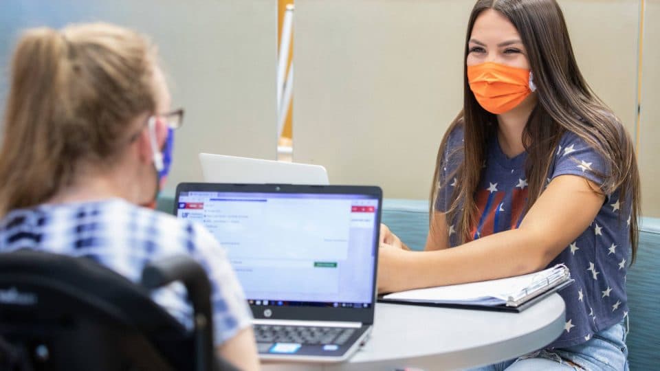 Students wearing masks interacting at a table with laptops and notebooks