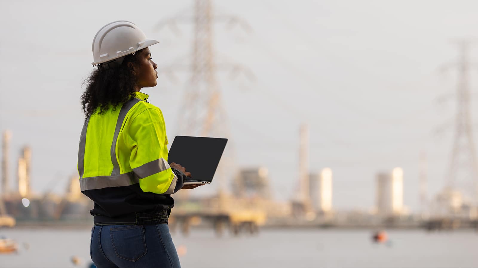 A female engineer wearing a hard hat and high-visibility jacket stands near an industrial site with power lines and refineries in the background, holding a laptop.