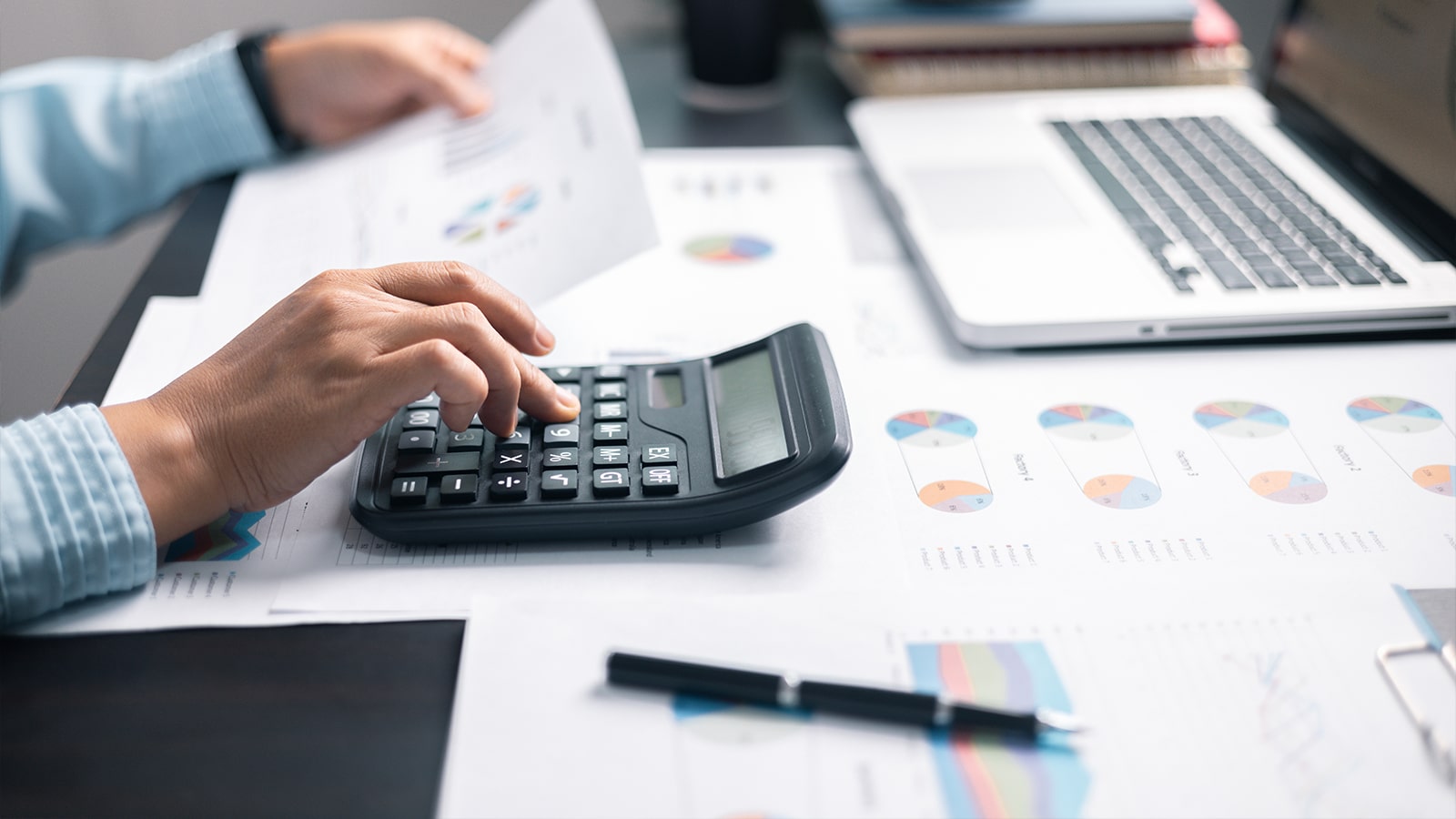 A person uses a calculator while reviewing printed financial charts and graphs, with a laptop and pen on the desk, representing financial analysis and investment strategy.