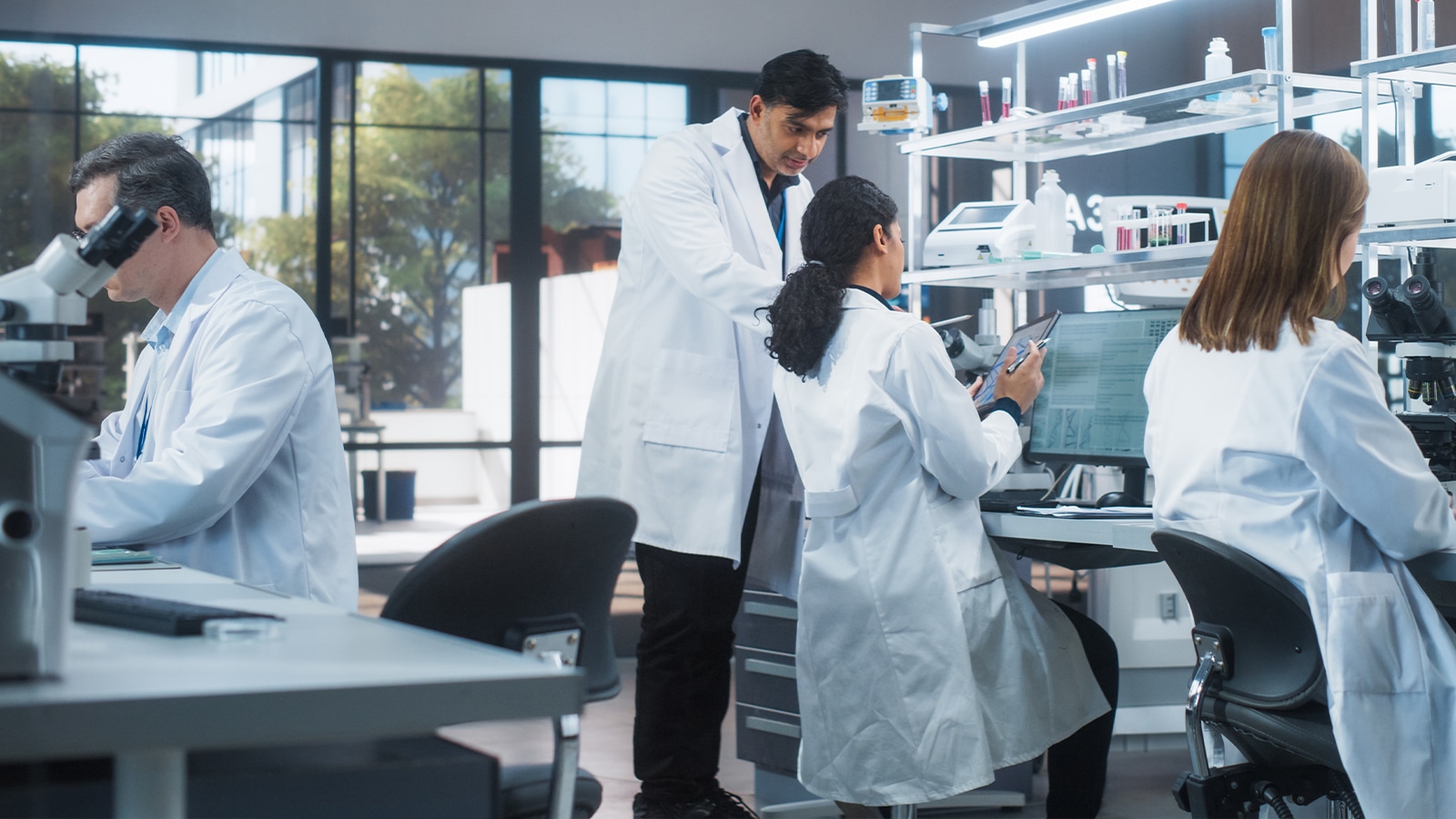 A group of scientists and medical researchers in lab coats work collaboratively in a modern laboratory, surrounded by microscopes, monitors, and lab equipment.