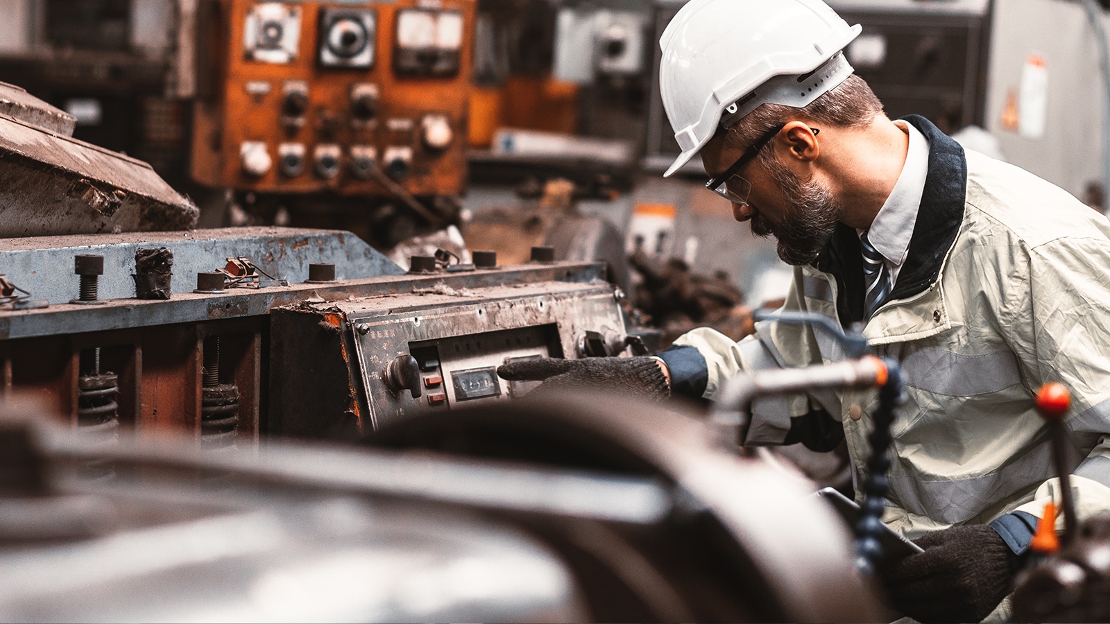 An industrial engineer in a hard hat and safety jacket operates machinery in a manufacturing or processing facility, surrounded by heavy equipment and control panels.