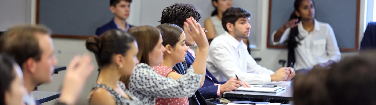 Graduate students in a classroom attentively listen and raise their hands to participate during an elective course session.