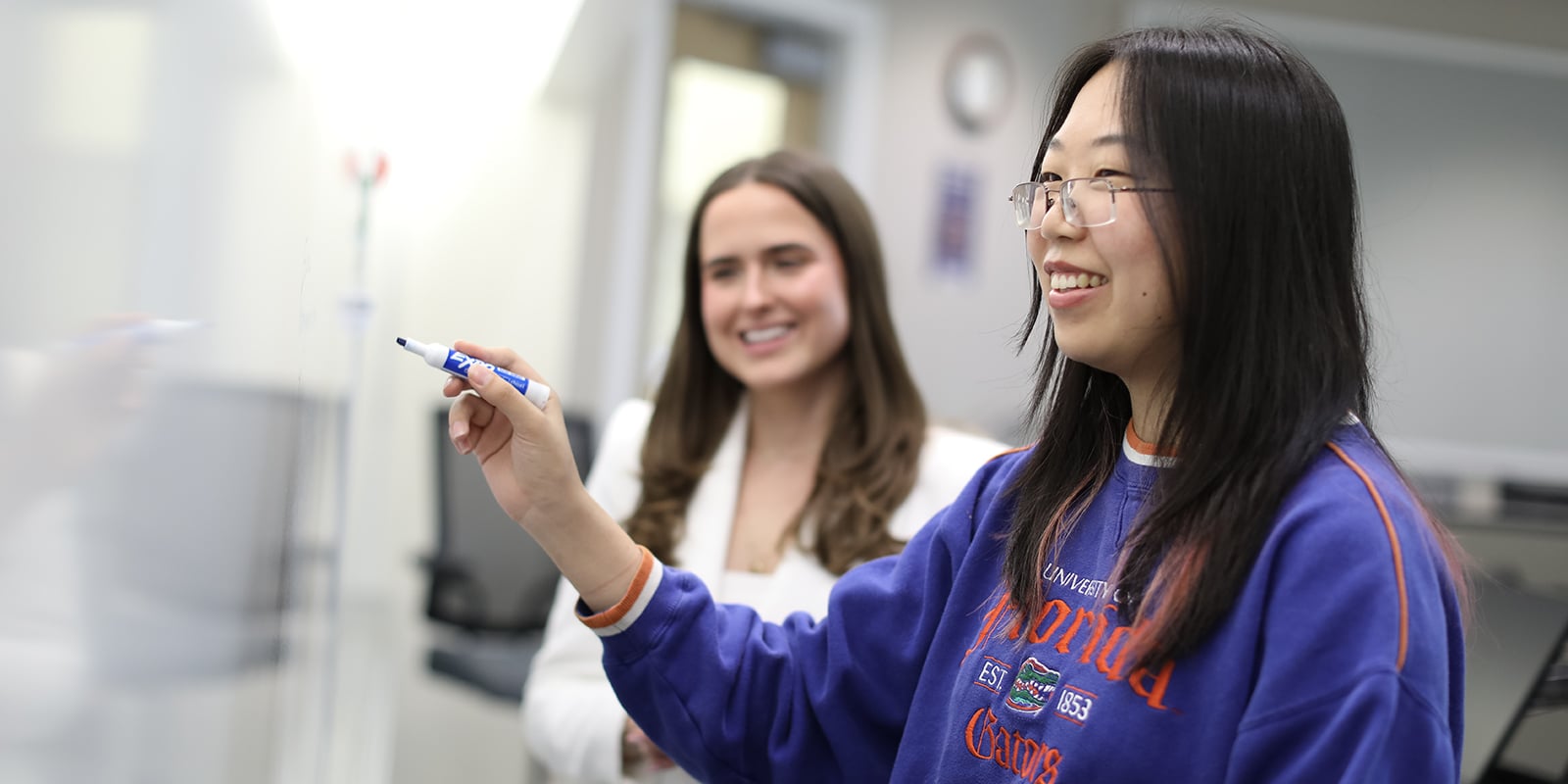 To fulfill UF Warrington's professional development requirements, a BS in Accounting student writes on a whiteboard for a problem-solving challenge while an instructor looks on