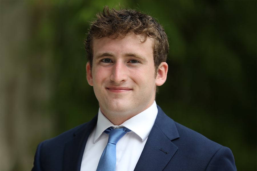A young man in a navy suit, white shirt, and light blue tie smiles confidently while standing outdoors with a blurred green background.