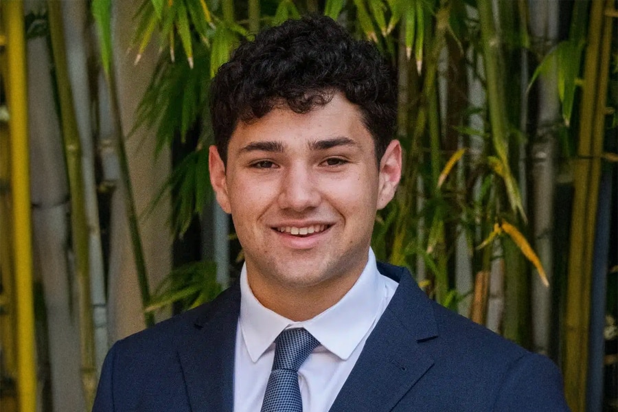 A young man in a dark blue suit, white shirt, and patterned tie smiles confidently in front of a backdrop of green bamboo stalks.