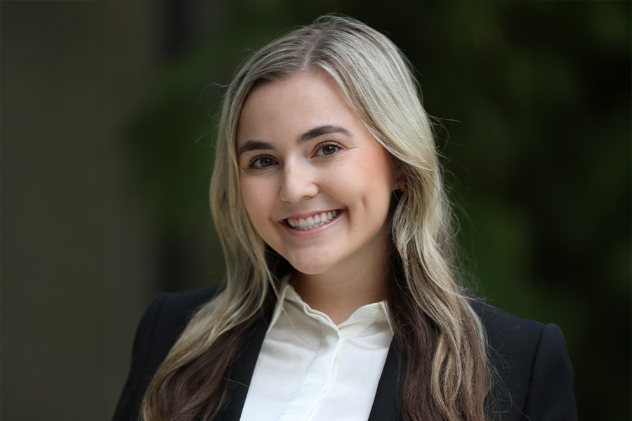 A young woman with long, wavy blonde and brown hair, dressed in a black blazer and white blouse, smiles warmly in an outdoor setting with a soft green background.