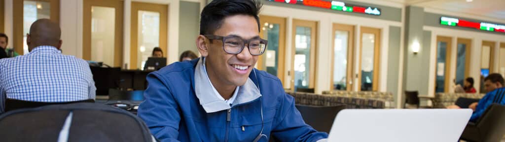 A Warrington College of Business undergraduate student, smiling and working on a laptop in a study area with digital stock tickers in the background, surrounded by other students.