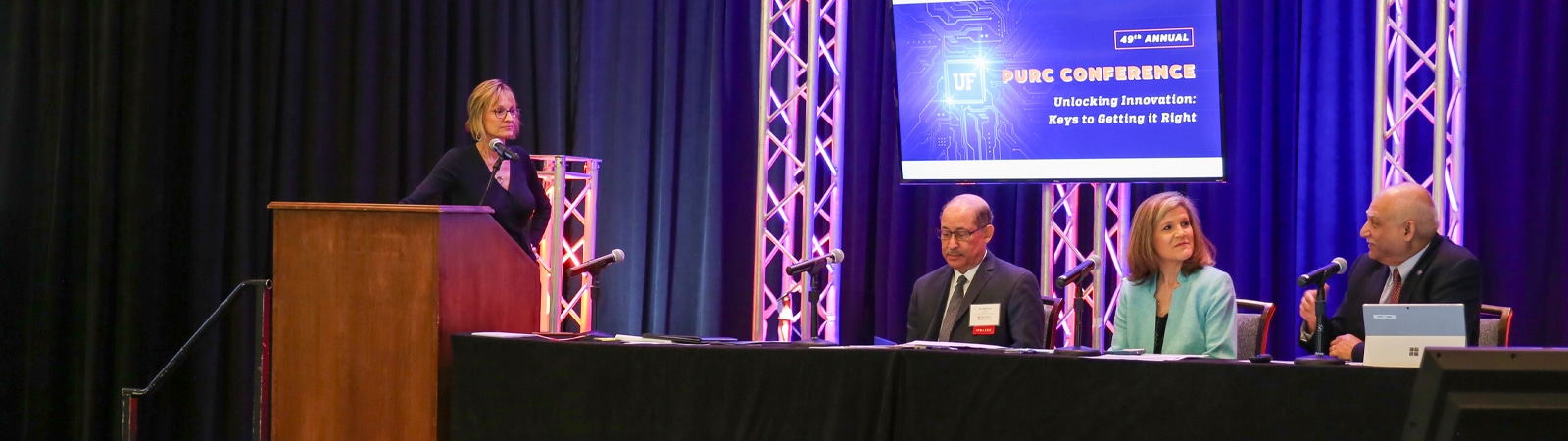 A panel of speakers and a moderator participate in a discussion at the 53rd Annual PURC Conference, with a digital display in the background highlighting the conference theme: "Unlocking Innovation: Keys to Getting it Right."