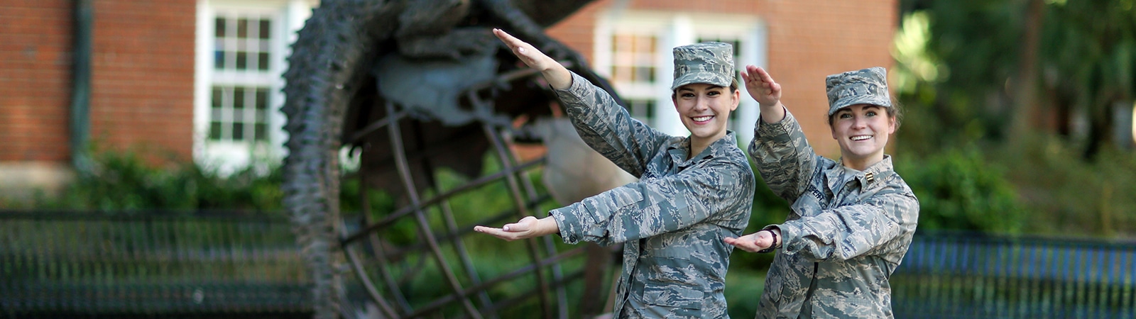 Two students wearing camouflage military uniforms, both enrolled in UF Warrington's BABA program, make Gator arms in front of the Gator statue