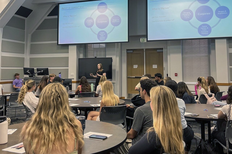 A female student leads a presentation to a room full of attentive undergraduate students seated at round tables in a classroom.