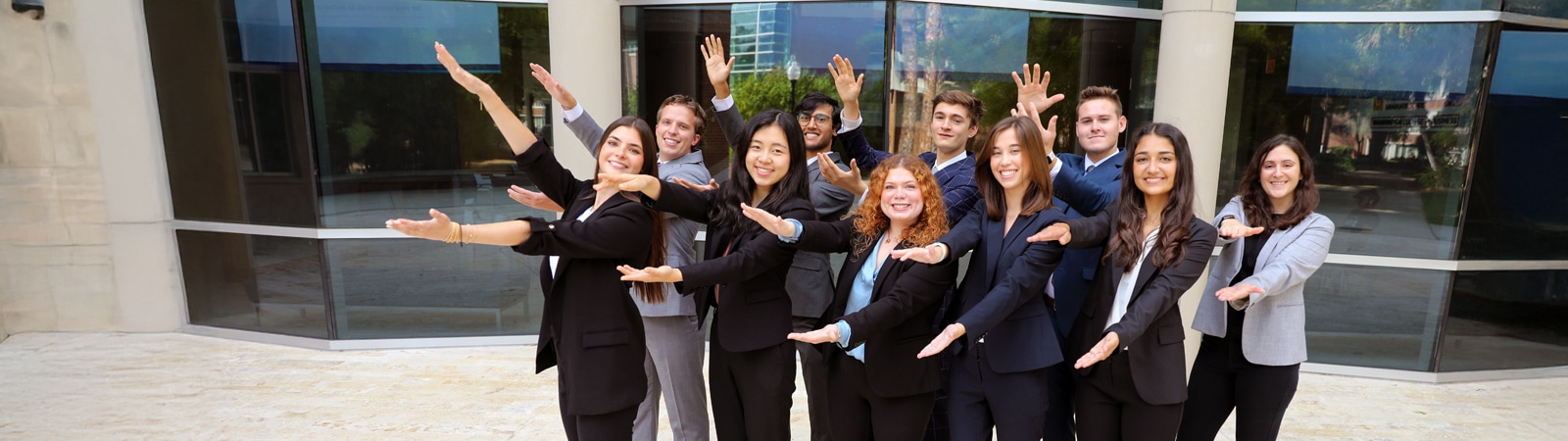 A group of business students in formal attire stand outside a modern glass building, smiling and posing enthusiastically with arms extended in alternating directions.