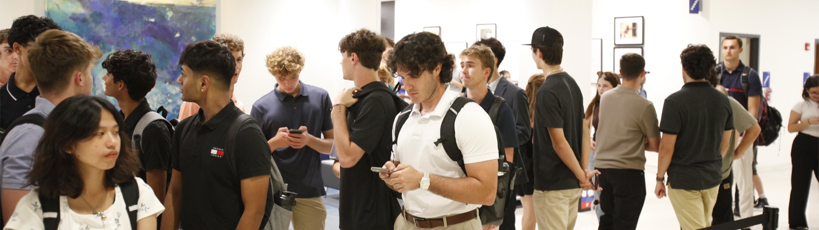 A diverse group of students stand in line and converse in a bright, modern hallway with backpacks and casual attire.