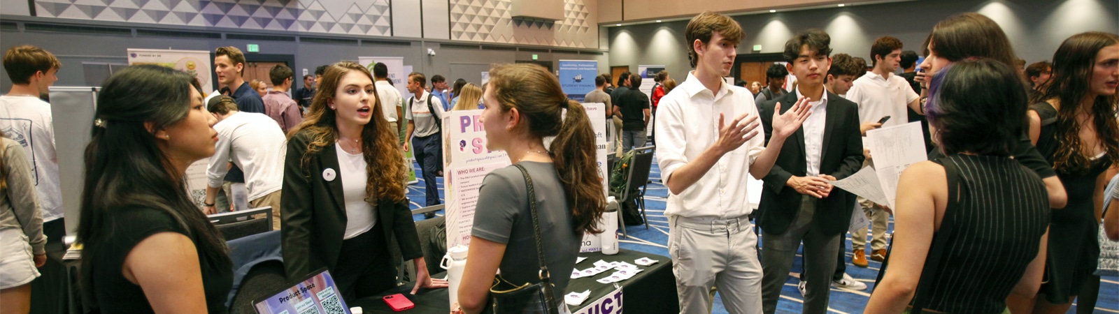 Students network and discuss at a busy career fair in a large indoor space with tables and displays.