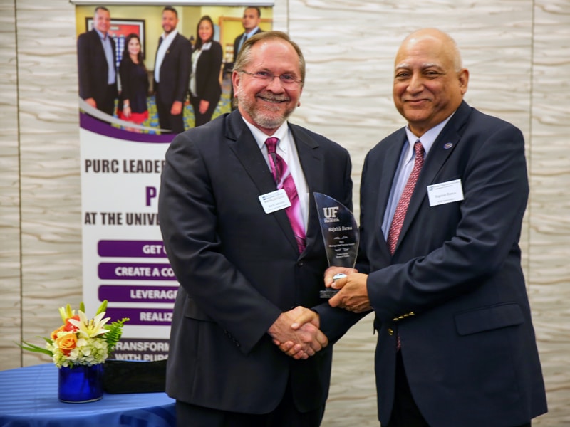 Two men in suits shake hands as one presents the other with the UF PURC Distinguished Service Award at the PURC Annual Conference, with a PURC Leadership Services banner and flower arrangement in the background.