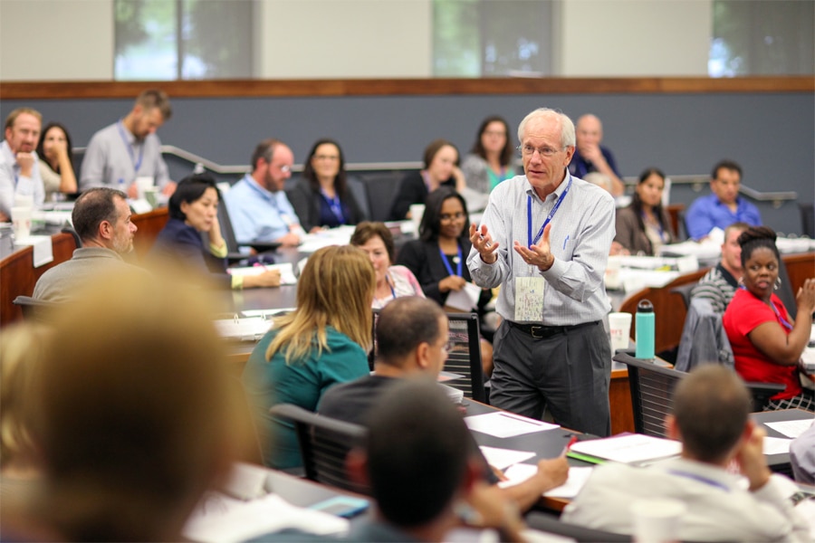 An older man with white hair stands in a classroom, actively engaging with a diverse group of seated participants who listen attentively and take notes.