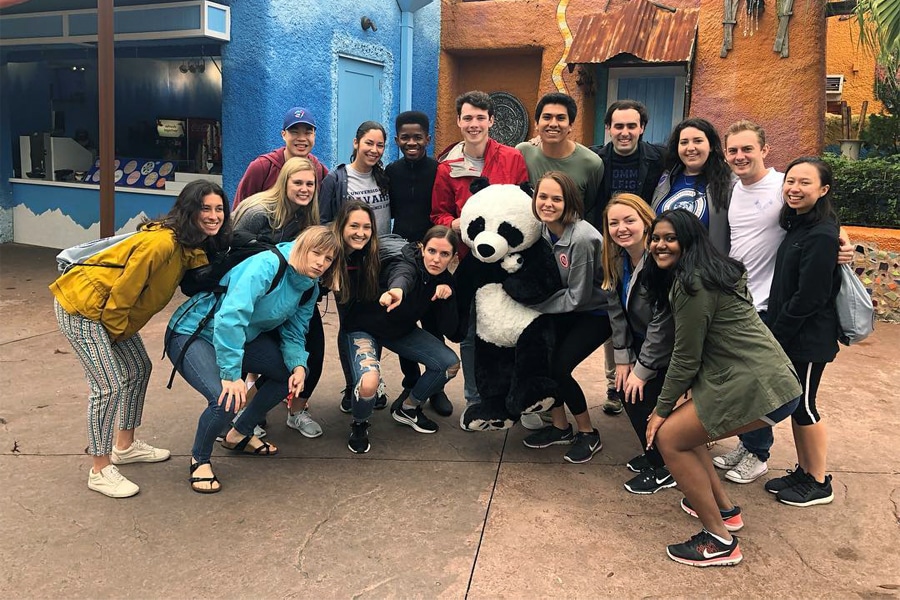 A diverse group of university students posing outdoors with a person in a panda costume, smiling and enjoying a day out at a colorful amusement park.