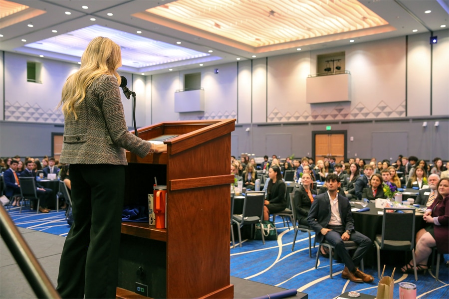 A woman speaks at a podium to a large audience seated at round tables in a conference hall with modern lighting and blue carpeting.