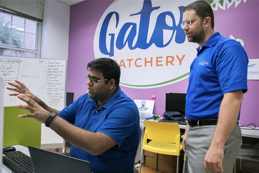 Two men in blue shirts, one seated and gesturing as he explains something, and one standing and listening, work together in the Gator Hatchery workspace at the University of Florida. A whiteboard with notes and a large sign reading “Gator Hatchery” can be seen in the background.