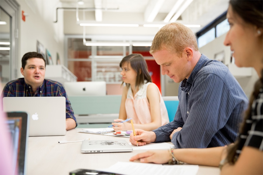 A group of students sit around a table in a bright, modern workspace, working on laptops and discussing ideas. One student is talking while others take notes, indicating active collaboration and brainstorming.
