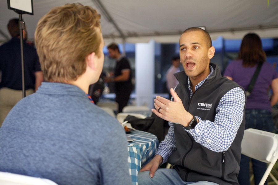 A man in a CENTENNIAL vest gestures as he speaks to a younger man seated across a table at an outdoor event.