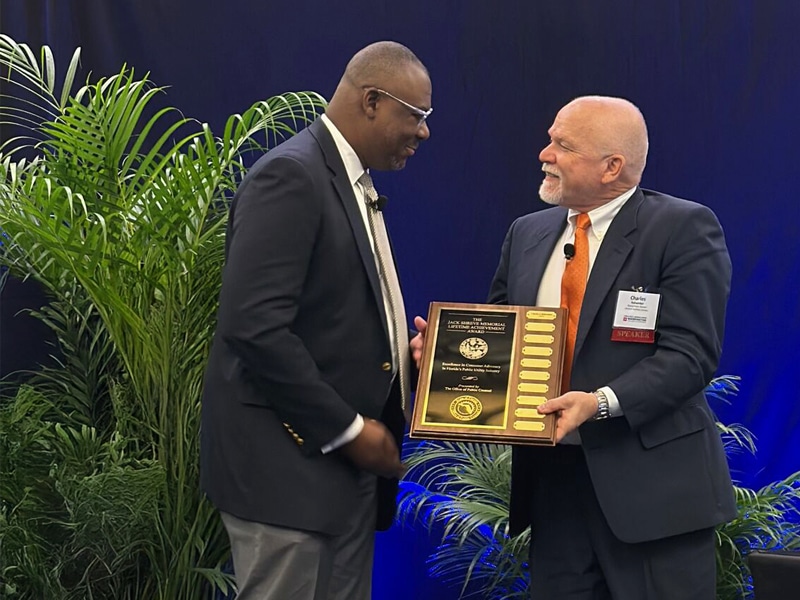 A man in a suit presents the Gerald L. Gunter Distinguished Service Award plaque to another man in a suit at the PURC Annual Conference, with lush greenery in the background.