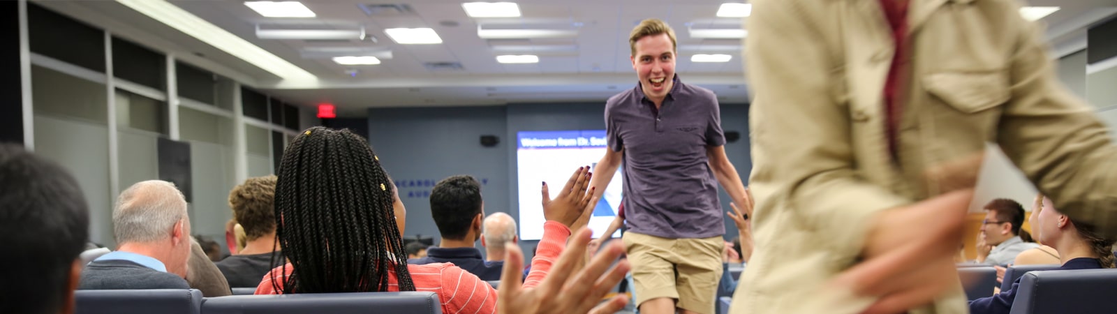 A student enthusiastically runs down an aisle, high-fiving audience members during an energetic moment at a business case competition event.