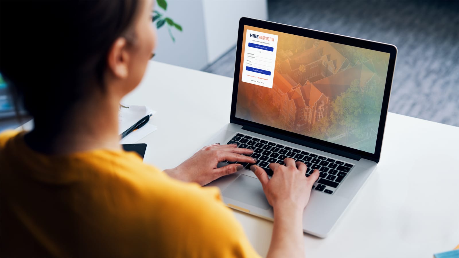 A person wearing a yellow sweater types on a laptop keyboard. The laptop screen shows the HIREWarrington login page, with options to log in using GatorLink or a student account. The background image on the screen features an aerial view of the University of Florida campus.