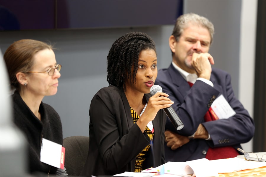 A woman speaks into a microphone while seated at a panel, with two colleagues listening attentively during a PURC International Advisory Council meeting.