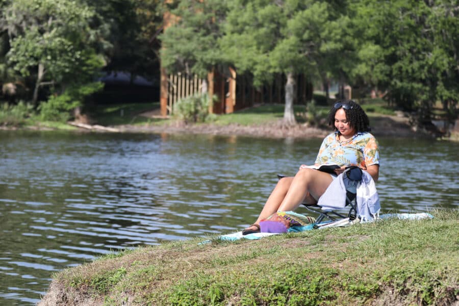 A young woman sits by a lake reading a book on a sunny day.