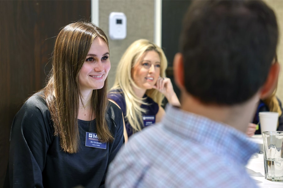 A young woman with a UF MBA name tag smiling attentively during a discussion, seated at a conference table with other students and professionals in a bright room.