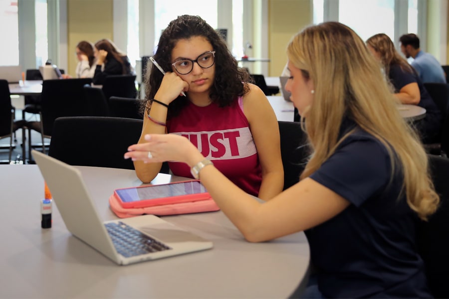 A young woman in glasses listens attentively while another woman gestures explaining something in front of a laptop in a study area.