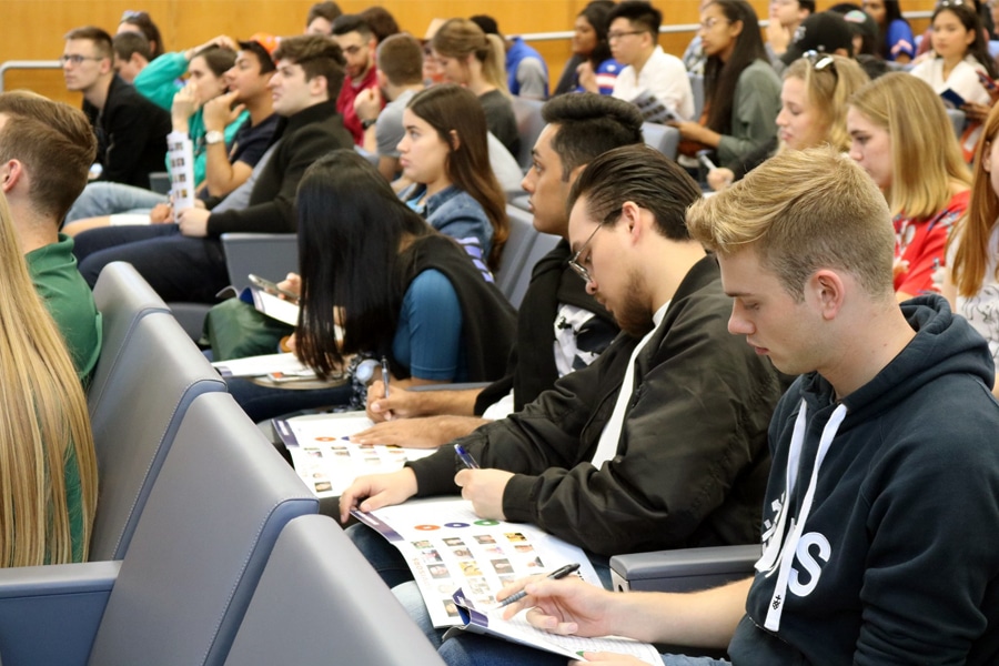 University students seated in rows in a lecture hall, focused on writing notes in booklets during a preparation session.