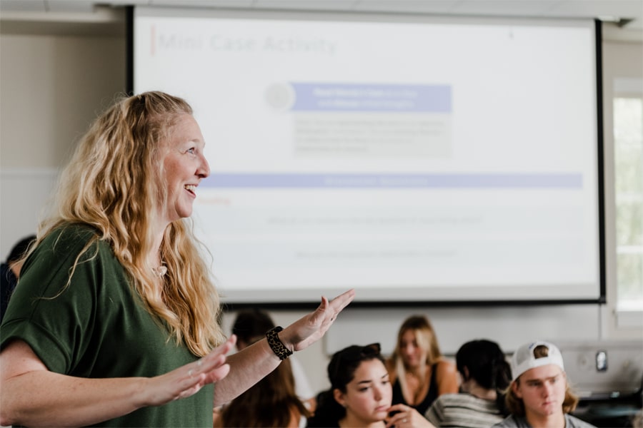 A female faculty member with long curly hair speaks animatedly in front of a classroom full of attentive students, with a presentation slide titled “Mini Case Activity” projected behind her.