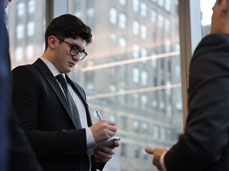 Young man in professional attire taking notes when speaking to a businessman.