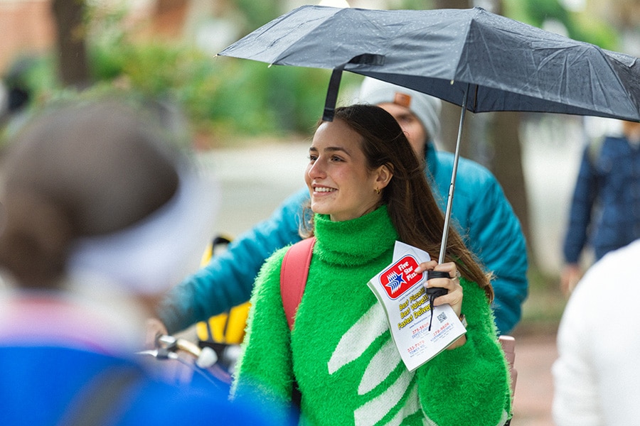 Young woman in a green sweater holding an umbrella and walking through a crowd on campus.
