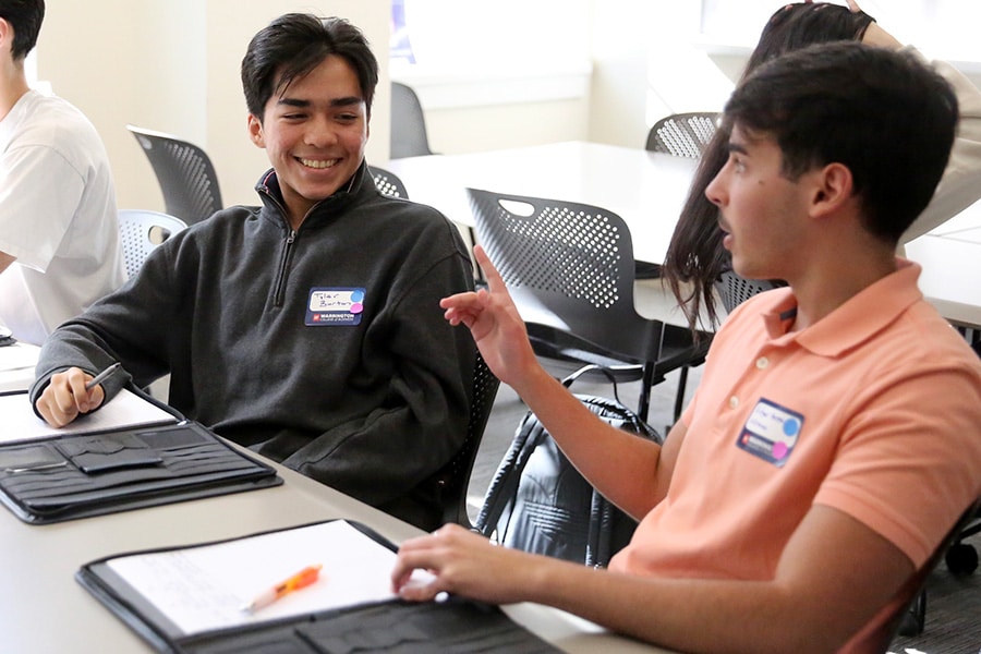 Two young men sit at a table with their notepads and conversing with each other.