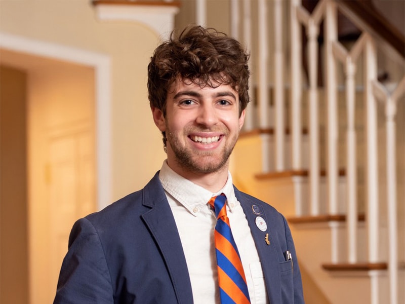 A young man with curly brown hair and a bright smile wears a blue blazer, a white shirt, and an orange-and-blue striped tie. He is standing in a well-lit home or office with a staircase in the background.