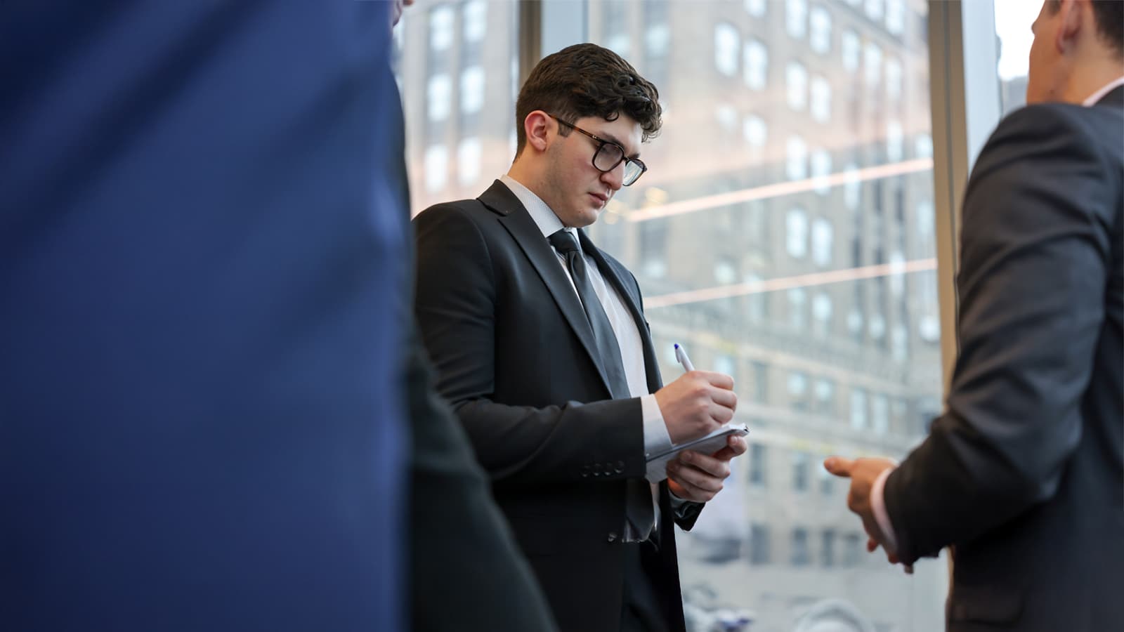 A young man in a black suit and glasses takes notes on a pad during a business meeting, with city buildings visible through a large window behind him.