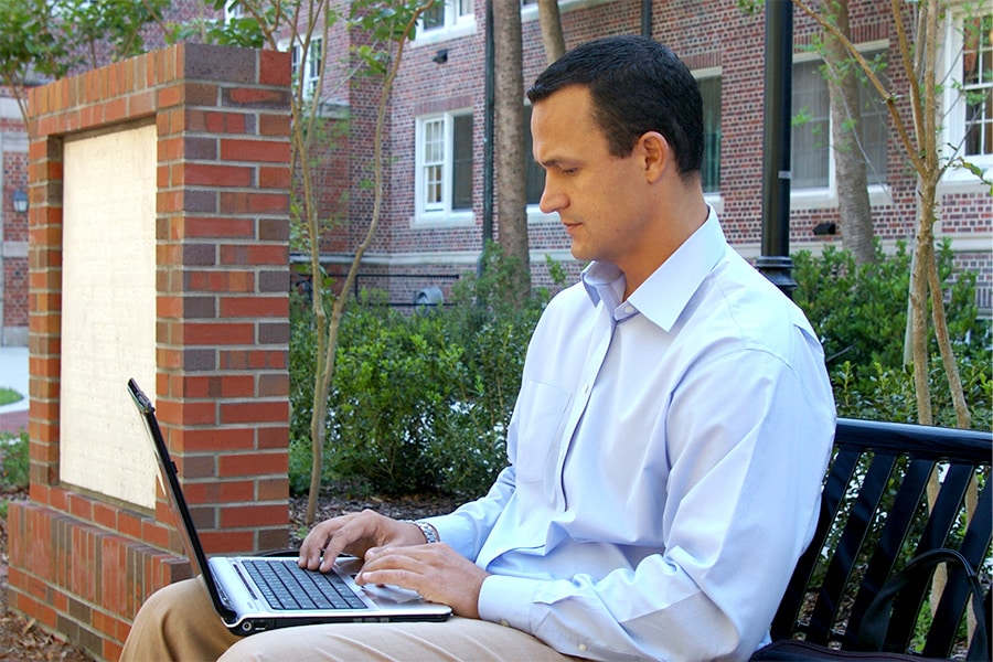 Man sitting outside on a bench with a laptop on his knees.