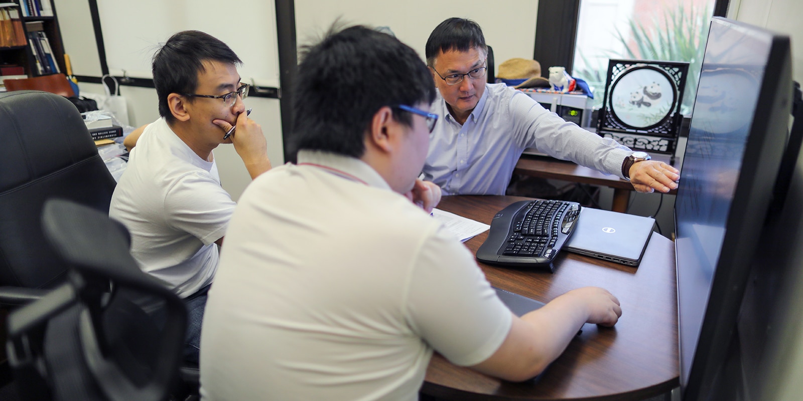 Three men sitting at a table around a monitor discussing research.