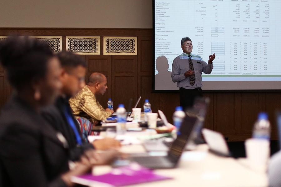 People sitting at a table with their laptops and notes looking towards a man instructing in front of a presentation.