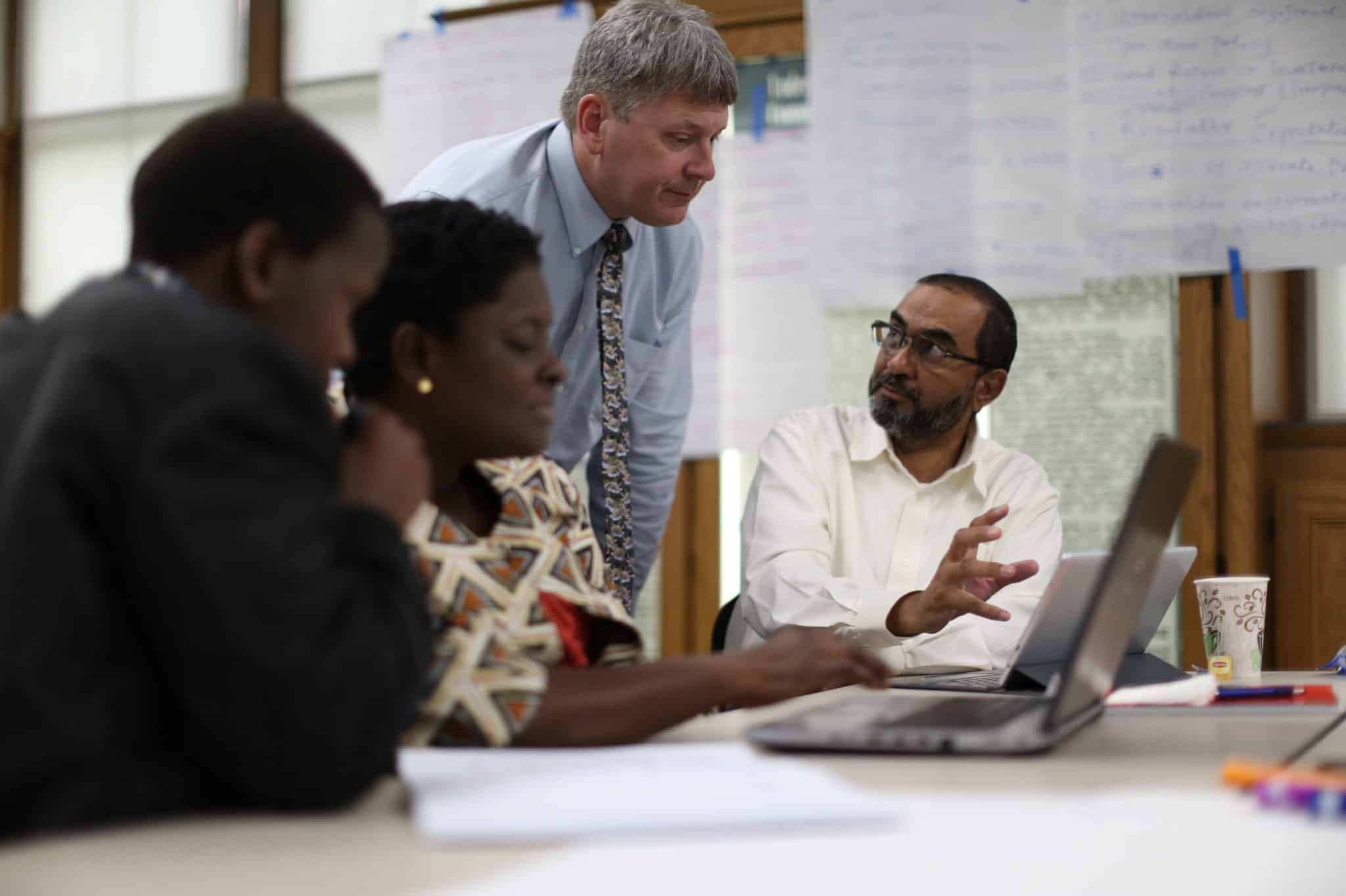 A man in a dress shirt and tie speaks with three adult learners in a classroom setting.