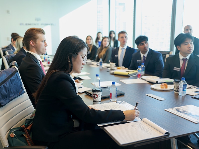 Students in professional attire sitting around a conference room table taking notes with the focus on a girl at the front of the table writing on her notepad.