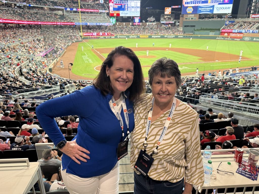 Two women stand together with a baseball field behind them. 