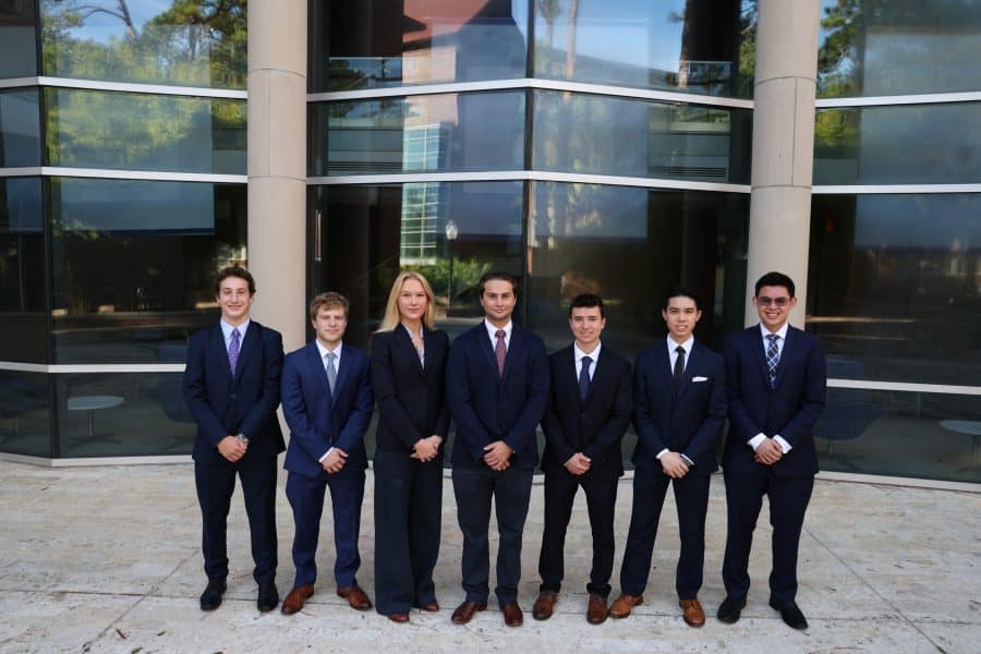 A group of seven students wearing suits pose for a photo in front of a large glass window.