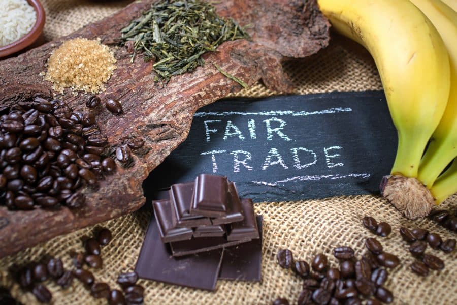 Assorted fair trade products including coffee beans, bananas, and chocolate displayed around a chalkboard sign that reads 'FAIR TRADE' on a burlap surface