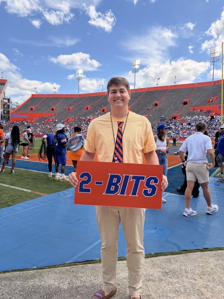 Blake Cox stands on the Ben Hill Griffin Stadium field holding an orange and blue sign that reads "2-Bits."